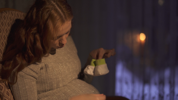 Raskaana oleva nainen, jolla on pitkät hiukset, istuu keinutuolissa pimeässä huoneessa. Lady leikkii pienillä vauvasukilla. Nainen jäljittelee pieniä askelia sukat vatsaan, video
  - Materiaali, video