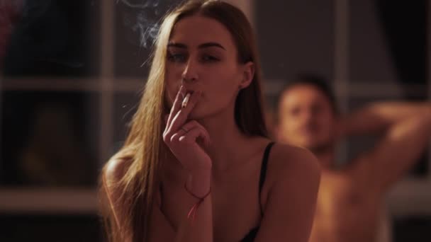Gyönyörű nő fehérneműben cigarettázik egy félig öltözött férfi előterében, aki az ágyban fekszik. A lány pihen, miután szeretkezett a sötét szobában. Az egészségtelen életmód fogalma - Felvétel, videó