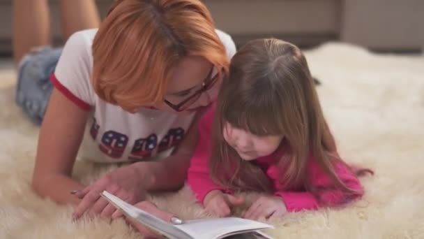 Beautiful mother and cute little daughter are lying on the carpet on the floor in the bedroom at home. Mom reads the girl a book. - Filmati, video