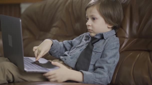 The little boy in a jeans jacket sitting on the leather sofa playing on the laptop. The leisure of modern child. Addiction to gadgets - Footage, Video