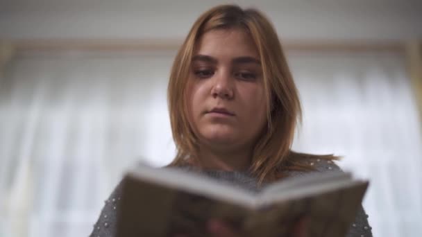 Plump girl reading book sitting at sofa in home. Chubby woman holding book in hands. Leisure of cute smart plus size lady at home - Materiał filmowy, wideo