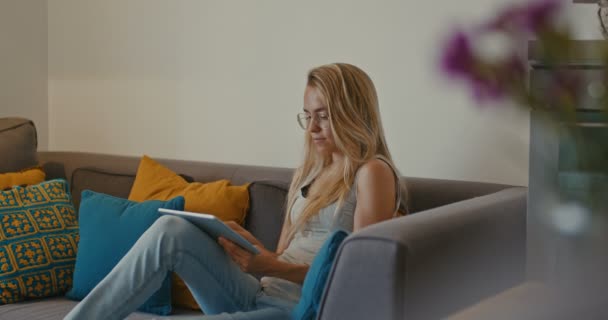Magas minőségű 4k 12bit felvételeit gyönyörű lány segítségével Tablet PC ül a kanapén otthon. Nő szörfözés web, szociális hálózatok, keres valamit az online boltban. Filmszerű megjelenés - Felvétel, videó