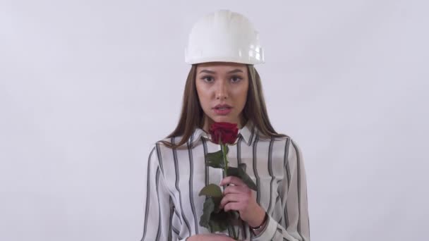 Young beautiful woman in construction white hat holding a red rose in her hands and very happy smelling flower. - Filmati, video