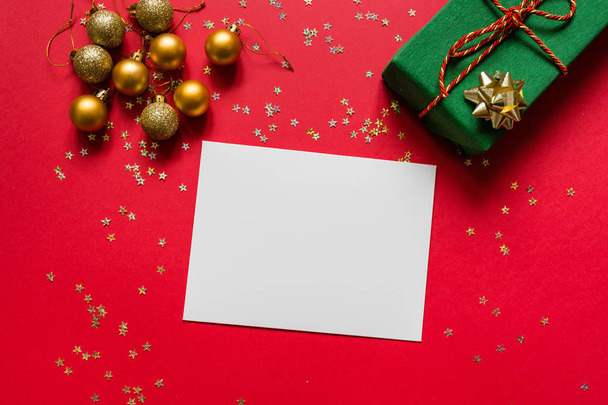 Christmas composition. White blank paper, wrapped gift, toys on red background with golden confetti. new year concept. Greeting card, xmas celebration 2020. Flat lay, top view, copy space, mockup - Φωτογραφία, εικόνα