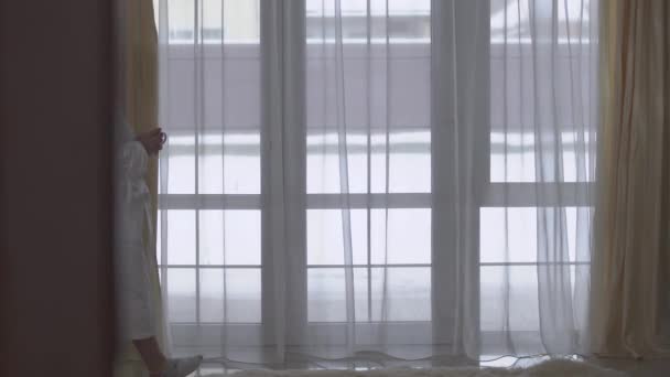 Plump girl in white long bathrobe walks to large floor-to-ceiling window with cup of coffee or tea. Lady straightening the tulle. Morning of happy young chubby woman - Filmmaterial, Video