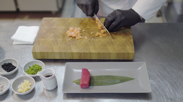 Mãos de chef no uniforme de restaurante branco que corta o pequeno peixe de salmão. Utensílios de cozinha, especiarias, pedaços de atum, molhos e outros ingredientes
. - Filmagem, Vídeo