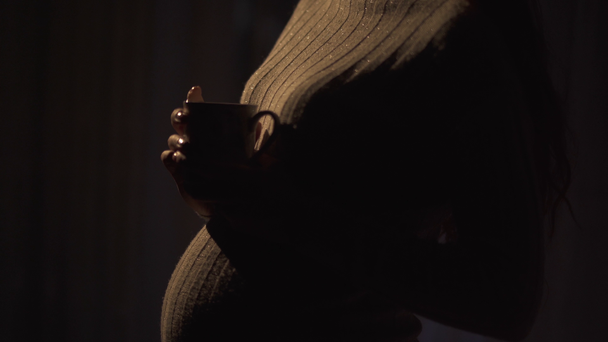 έγκυος κοπέλα στέκεται σε ένα ζεστό πουλόβερ σε ένα σκοτεινό δωμάτιο το βράδυ κρατώντας ένα φλιτζάνι ζεστό τσάι, βίντεο  - Πλάνα, βίντεο