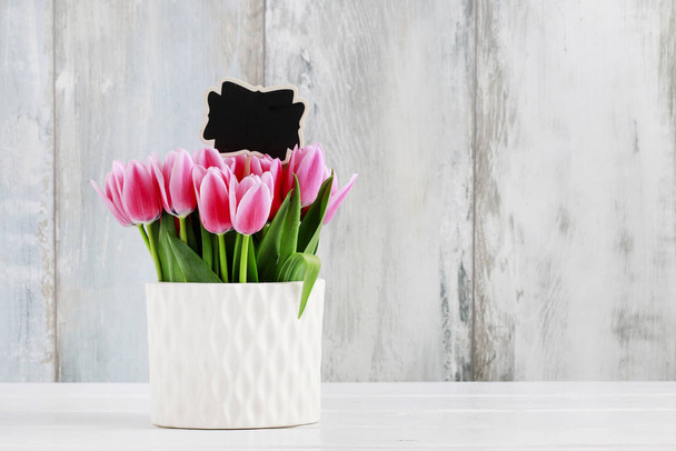 Pink tulips in white ceramic vase, wooden wall background.  - Foto, afbeelding