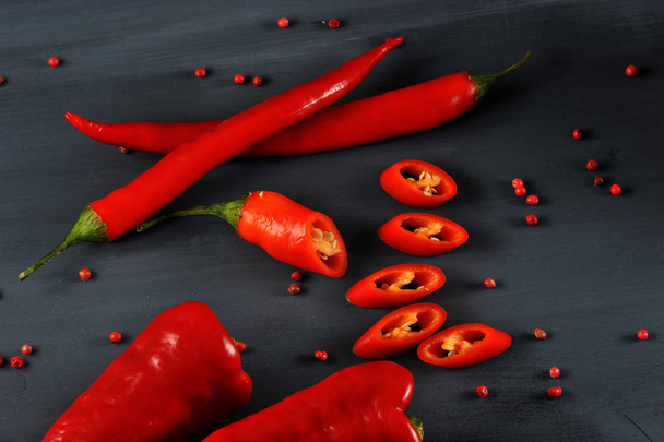 Spicy cayenne pepper on a dark background. In the frame is also a variety of sweet pepper Ramiro. One hot chili pepper is cut into mugs. Pears of pink pepper are scattered on the surface. Close-up. - 写真・画像