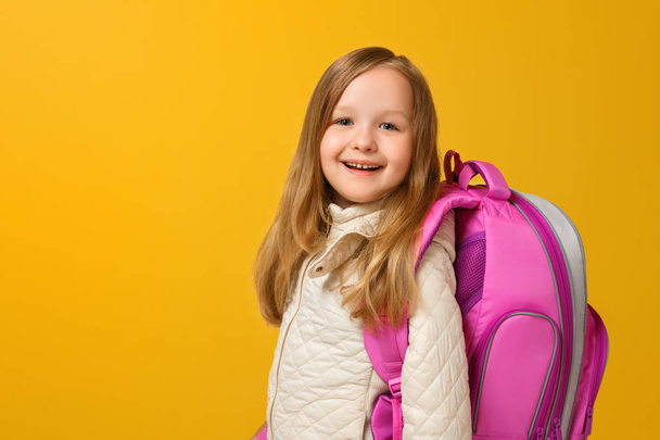 Portrait of a little girl schoolgirl in a jacket with a backpack on a yellow background. Back to school. The concept of education. Copy space - Φωτογραφία, εικόνα