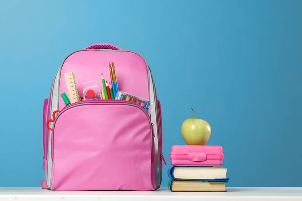 Student set. Pink backpack with stationery, a stack of books, a lunchbox, an apple on the table on a blue background. Back to school. - Zdjęcie, obraz