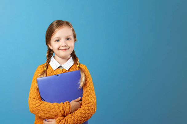 Closeup portrait of a little girl schoolgirl. Pretty child in a yellow sweater holds a purple folder for papers on a blue background. Copy space - Zdjęcie, obraz