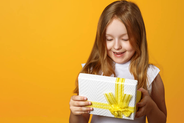 Portrait of a happy little girl opening a gift box with a gift on yellow background - Fotó, kép