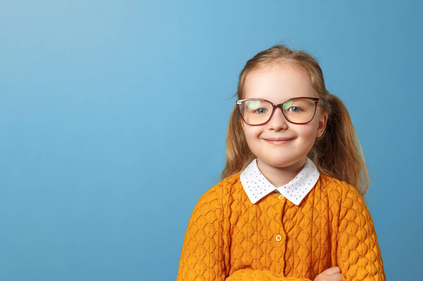 Closeup portrait of little girl schoolgirl with glasses. Pretty child in a yellow sweater on a blue background. Copy space - Fotografie, Obrázek