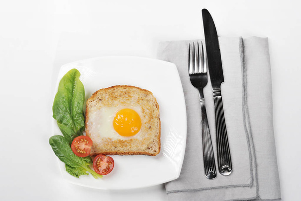 Toast with fried egg on a white plate. On a plate, spinach leaves and cherry tomatoes. Next to the dish there is a napkin and cutlery. White background. Close-up. View from above. - Foto, imagen
