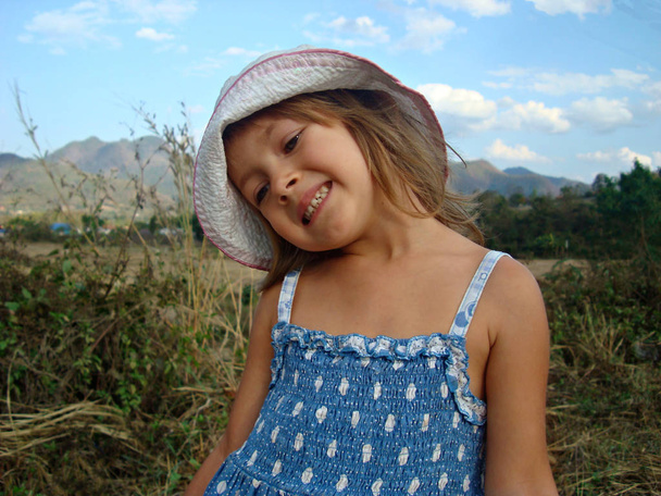 Egy kislány, kék ruhában, és egy rózsaszín sapka álmodosan és örömmel ellen kék eget, fehér felhők és dombok - Fotó, kép