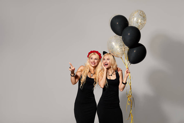 portrait of two happy young women tweens in black witch halloween costumes with balloons on party over white background. the concept of Halloween . funny faces. Showing hands to left, space for text. - Photo, Image