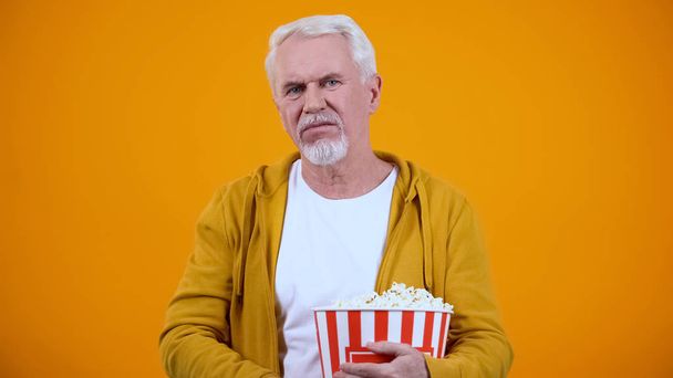 Starší muž s popovou kukuřicí rozrušený nudným filmem v kině, žluté pozadí - Fotografie, Obrázek