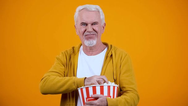 Old man displeased with uninteresting movie, holding popcorn, yellow background - Zdjęcie, obraz