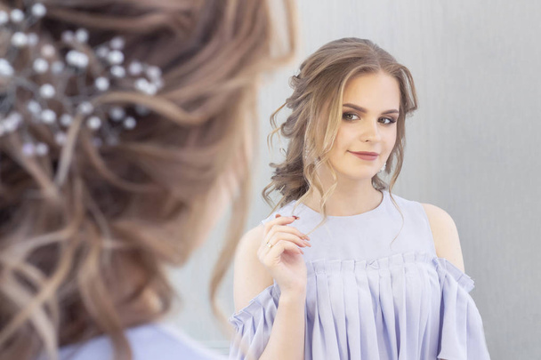 schöne Mädchen mit einer Hochzeitsfrisur sieht sich selbst im Spiegel, Porträt eines jungen Mädchens. Schönes Make-up. Schönheitssalon - Foto, Bild