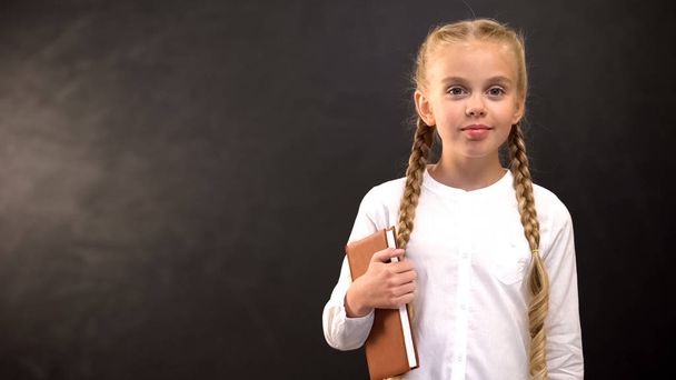 Pretty girl with book looking at camera against blackboard background at school - Фото, изображение