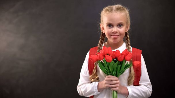 Pretty female first-grader holding tulips bouquet against blackboard background - Foto, imagen