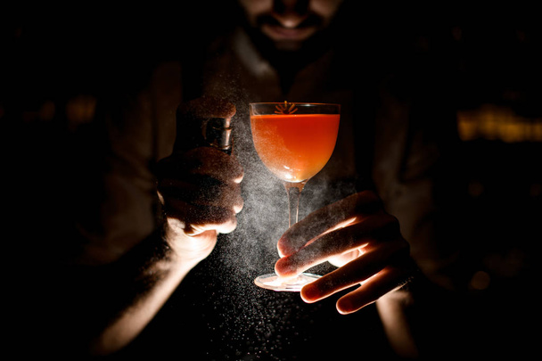Baarimikko tarjoilee ruskeaa oranssia cocktailia suihkuttaen sitä pimeässä
 - Valokuva, kuva