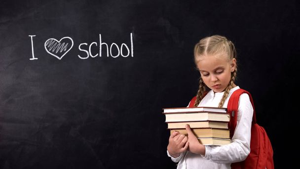 Sad girl with books standing against I love school phrase written on blackboard - Фото, зображення