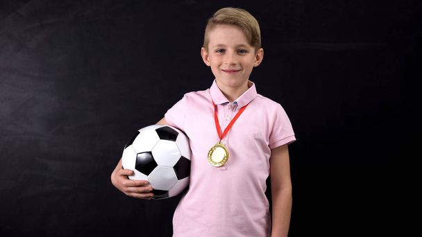 Αγόρι με μπάλα και μετάλλιο που στέκεται κοντά στον μαυροπίνακα, νικητής του αγώνα ποδοσφαίρου - Φωτογραφία, εικόνα