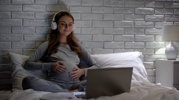 Gülümseyen hamile kadın dizüstü bilgisayarda film izlerken ve göbek tutarak, bekliyor - Fotoğraf, Görsel
