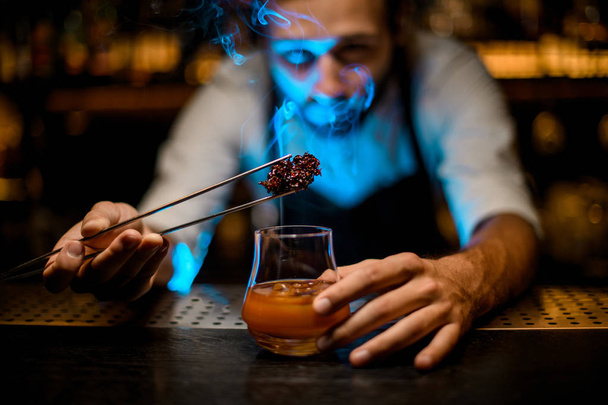 Attractive bartender adding chilled melting caramel with twezzers to the cocktail glass with ice cubes under blue lights - Фото, изображение