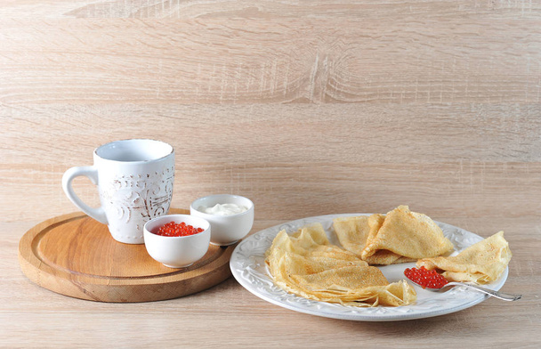 Pancakes on a plate and a teaspoon with caviar. Next to the wooden tray. On the tray there is a cup of tea, bowls with red caviar and sour cream. Light wooden background. Close-up. - Foto, Imagen
