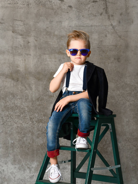 Cute stylish boy sitting on chair near concrete wall - Photo, image
