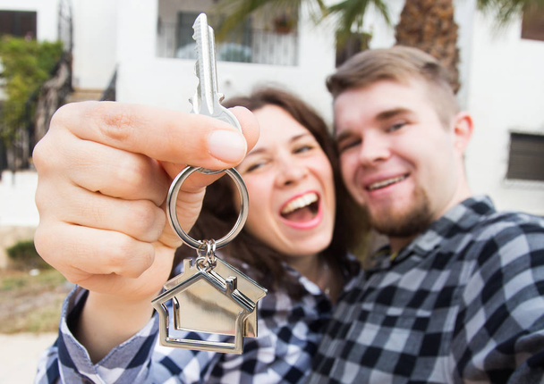 Το κατάλυμα, ακίνητα και διαμέρισμα έννοια - Happy αστεία νεαρό ζευγάρι που δείχνει μια κλειδιά του νέου σπιτιού τους - Φωτογραφία, εικόνα