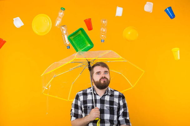 ゴミ、プラスチックリサイクル、汚染、環境コンセプトの問題 - 悲しい男は黄色の背景に傘でゴミの下に立っています - 写真・画像
