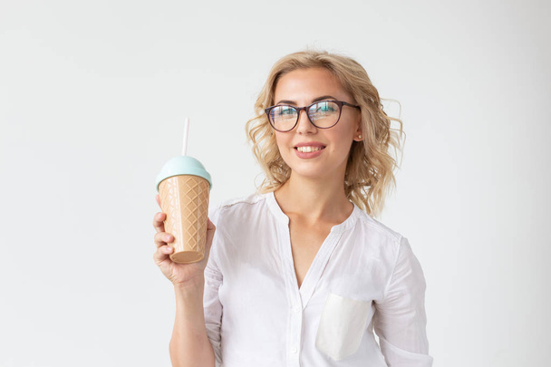 Krásná pozitivní mladá dívka v bílé košili má v rukou koktejl, který pózuje pro bílé pozadí. Pojetí dezertů a koktejlů. - Fotografie, Obrázek