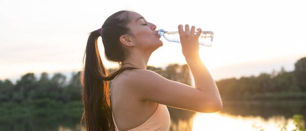 Hermosa atleta de fitness mujer beber agua después de hacer ejercicio en la puesta del sol noche verano en la playa retrato al aire libre
 - Foto, Imagen