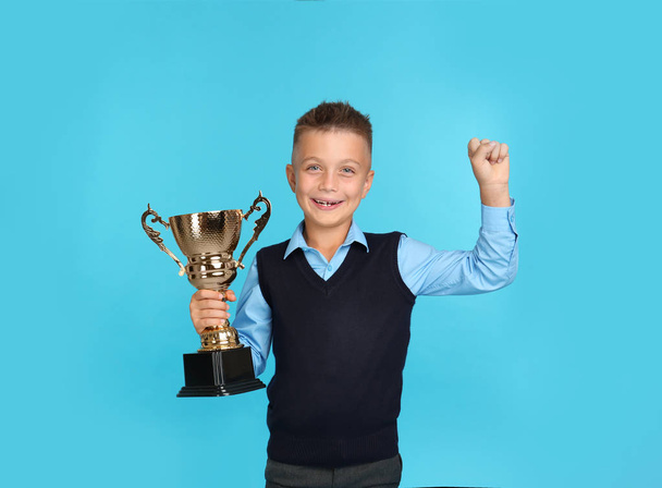 Happy boy in school uniform with golden winning cup on blue background. Space for text - Φωτογραφία, εικόνα