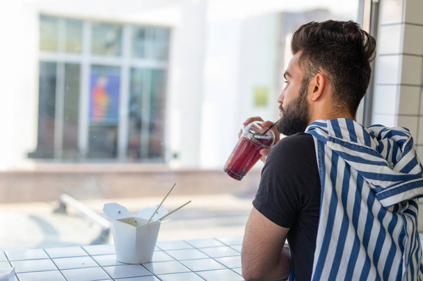 Позитивный молодой стильный парень пьет сок в кафе во время перерыва на работе. Концепция отдыха и здоровых закусок
. - Фото, изображение