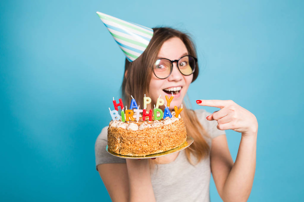 Encantadora joven estudiante loca alegre en sombrero de papel de felicitación sosteniendo un pastel de cumpleaños feliz en sus manos de pie sobre un fondo azul. Espacio publicitario
. - Foto, Imagen