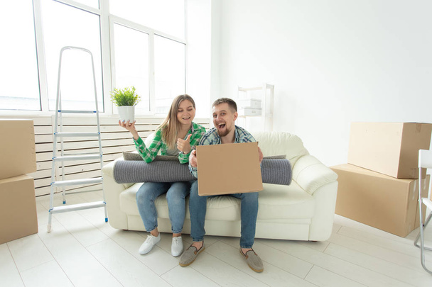 Χαρούμενο χαρούμενο νεαρό ζευγάρι γοητευτικό κορίτσι και όμορφος άνθρωπος κρατώντας ένα κουτί με τα πράγματα και μια κατσαρόλα με ένα φυτό, ενώ κινείται σε ένα νέο διαμέρισμα. Σχέδιο οικιακής σίτισης και ενυπόθηκης πίστης. - Φωτογραφία, εικόνα