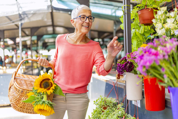 Attractive mature woman shopping in an outdoors fresh flowers market stall, buying and picking from a large variety of colorful floral bouquets during a sunny day in the city. - Foto, Imagem