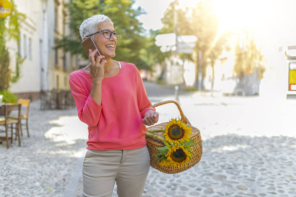 Elegant mature woman using smartphone while walking outdoors in city. Concept of business life on retirement. Happy senior woman walking along street and calling on smartphone. - Valokuva, kuva
