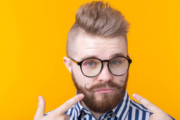 Charming confident young fashion hipster man with glasses and a beard shows on himself posing over a yellow background. Place for advertising. The concept of self-confidence and success. - Photo, image