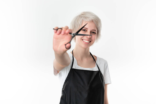 Pretty young hairdresser in white tee and black apron holding black scissors close to face ready for work standing isolated over white background. Wellness and beauty industry and professional career. - Photo, Image