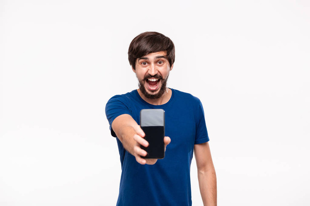 Un bell'uomo bruno con una camicia blu con barba e baffi che mostra lo schermo dello smartphone sulla fotocamera in piedi isolato su sfondo bianco. Emozione e gesto di sorpresa. Posto per annuncio
. - Foto, immagini