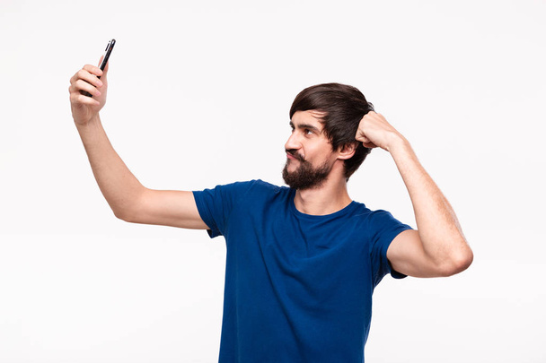 Beyaz arka plan üzerinde izole ayakta kas gösteren bir akıllı telefon ile selfie yapma bıyıklı yakışıklı brunet sakallı adam. - Fotoğraf, Görsel