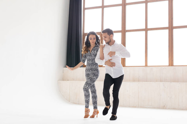 Молодая пара танцует социальные латинские танцы бачата, меренге, сальса. Две элегантные позы в танцевальном классе с большим окном
 - Фото, изображение