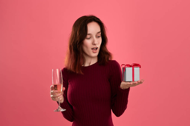 Excited pretty brunette girl holding present box with a ribbon in one hand and glass of champagne wine standing isolated on a dark pink background. Celebrating special event. - 写真・画像