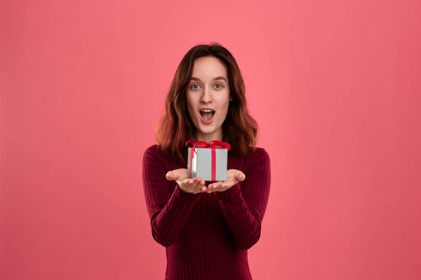 Excited pretty brunette girl holding present box with a ribbon with two hands standing isolated on a dark pink background and smiling at the camera. Celebrating special event. - Photo, Image